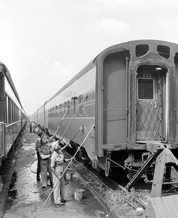 Three workers in coveralls use long sticks to clean the outside of a train car in an outdoor train yard