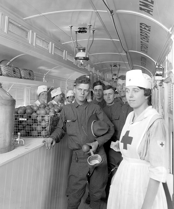 A line of young men in uniform surrounded by nurses inside a narrow train car. A bin of apples sits on the counter.