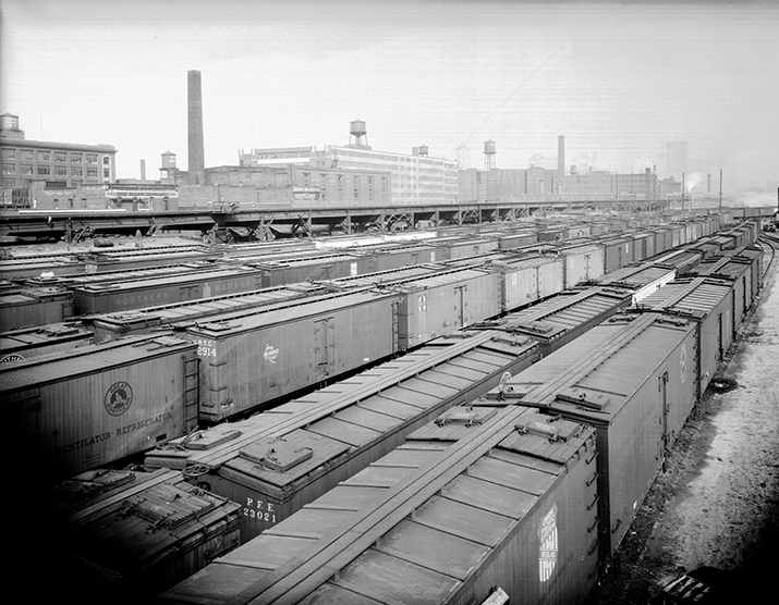 An overhead view of a railyard full of trains with industrial buildings in the background. Train cars labelled with text such as "union pacific system: overland" and "Great northern refrigerator"