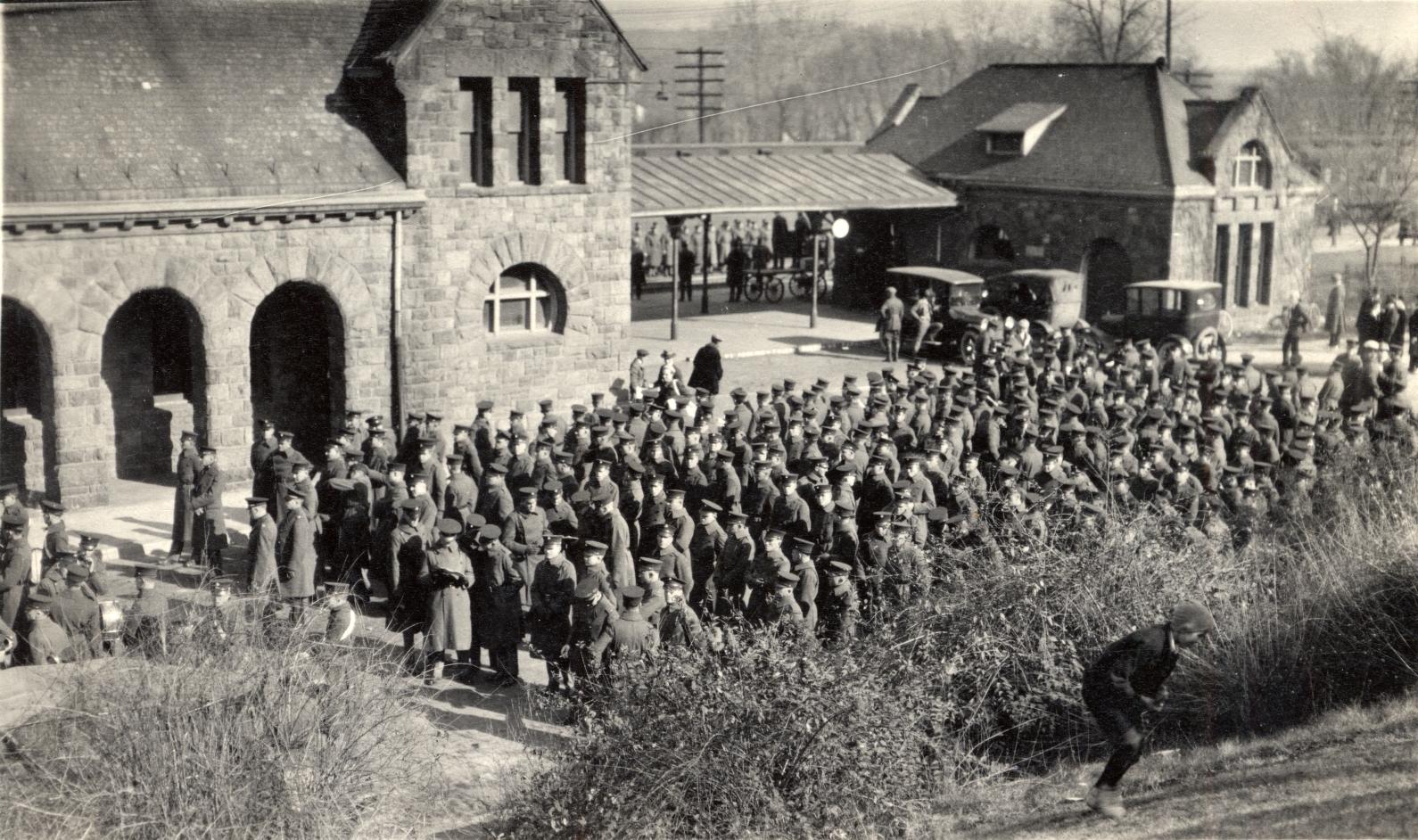 Roughly 100 soldiers in uniforms line up outside a stone railroad station