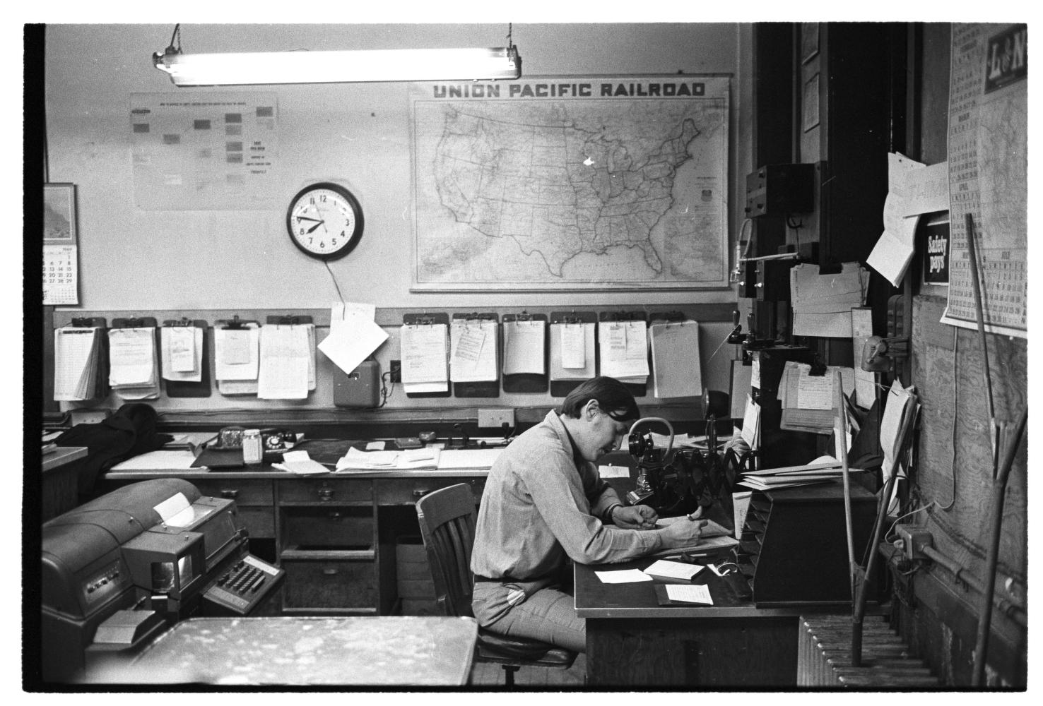 A man leans over a desk in a room full of clocks, maps, clipboards, and other equipment