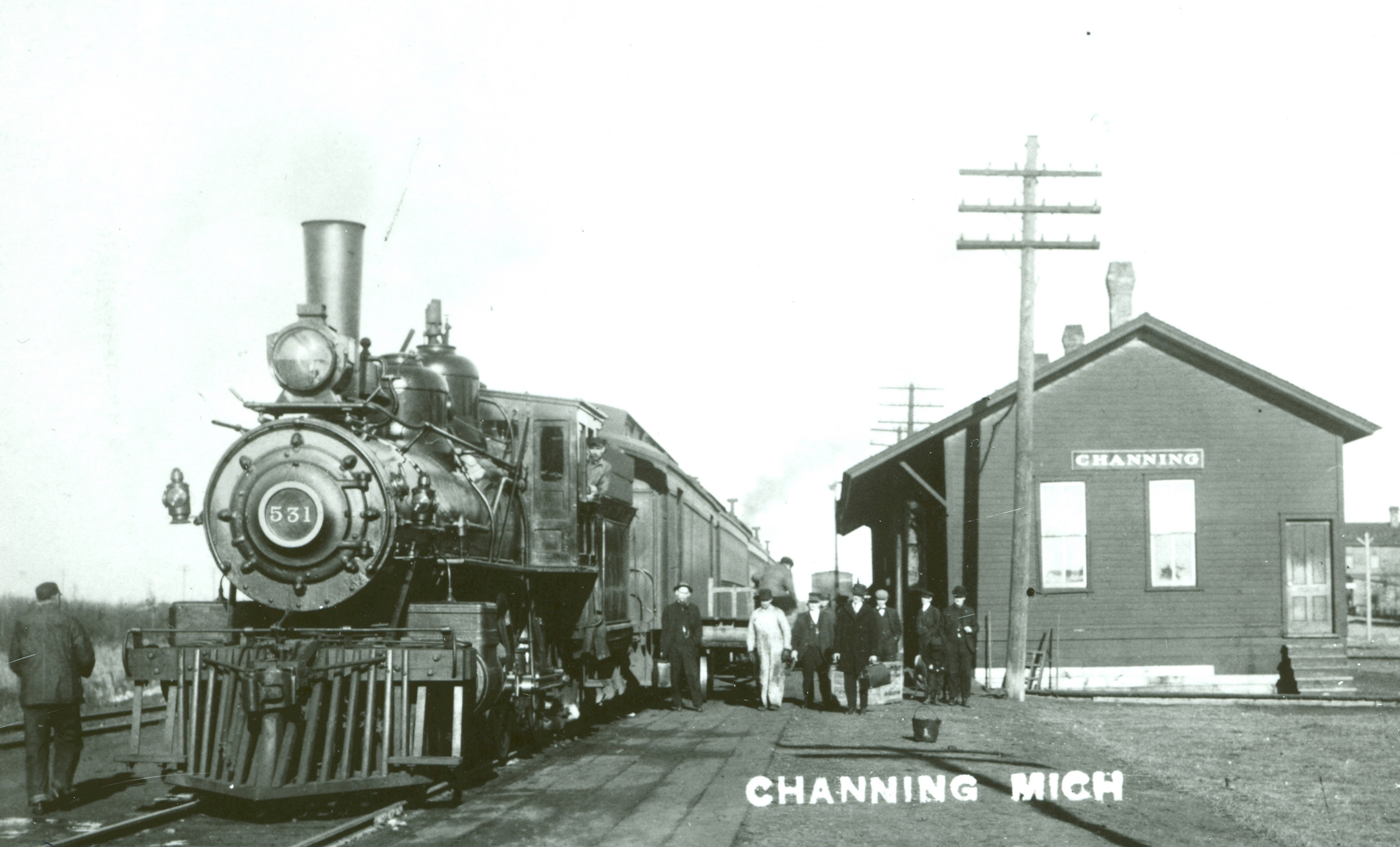 Passengers in hats and jackets between a small depot marked "Channing" and a locomotive and train marked 531.