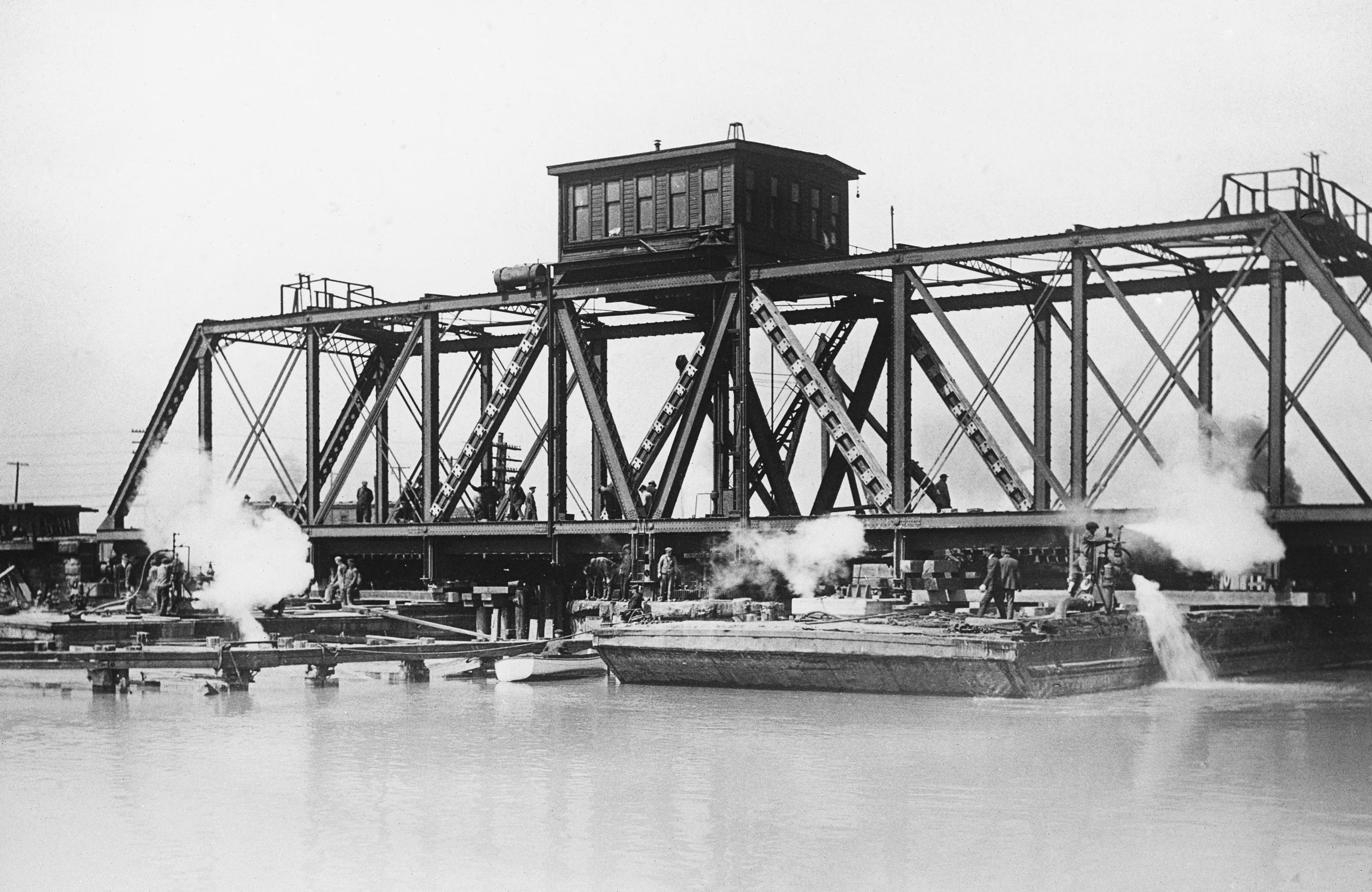 A metal frame railroad bridge surrounded by boats emitting steam